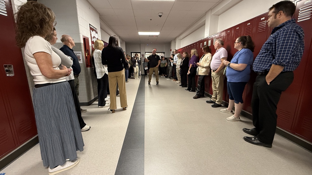 Several teachers sitting in a hallway up against lockers listening to a man speak