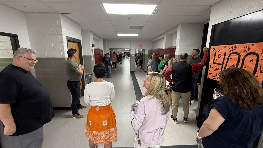 Several teachers sitting in a hallway up against lockers listening to a man speak