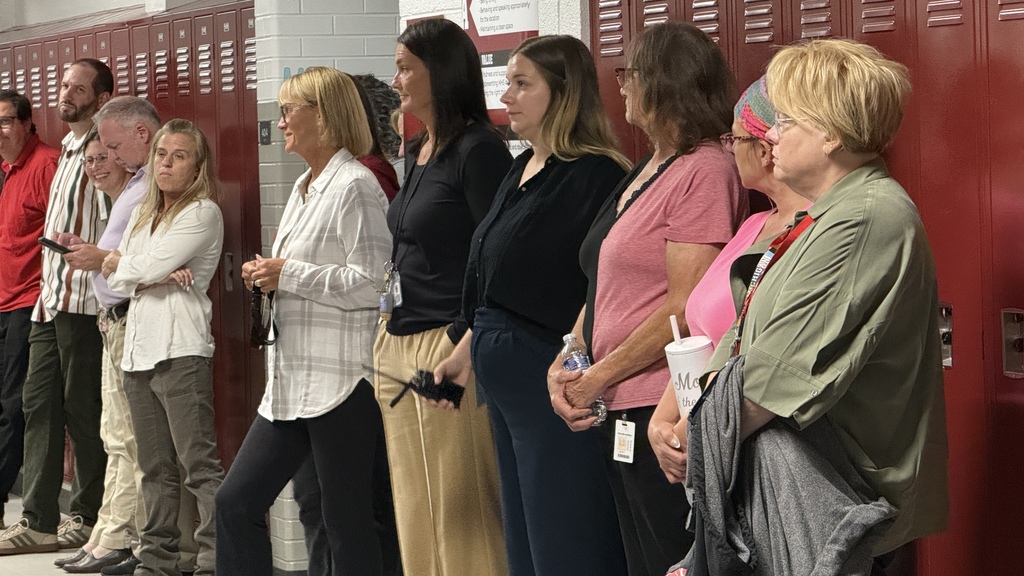 Several teachers standing up against lockers listening to a speaker 