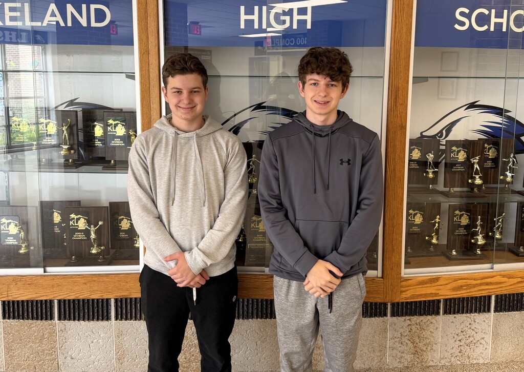 Two males students standing in front of a school trophy case smiling and posing for a photo
