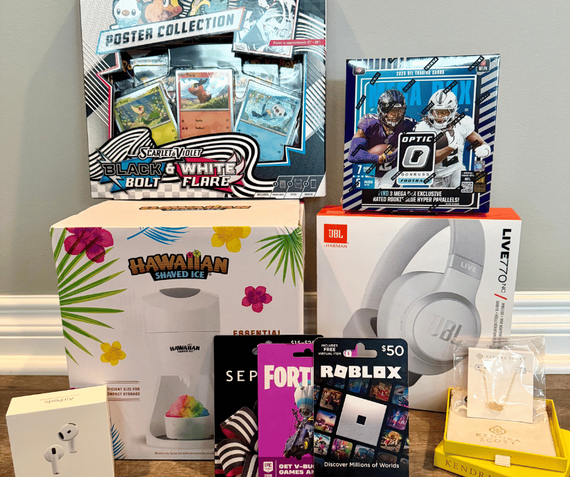 A collection of raffle prizes neatly displayed on a wooden surface:  Large Items: A box for a Hawaiian Shaved Ice machine and a box for JBL Live 770NC wireless headphones.  Card Sets: A Pokémon Scarlet & Violet "Black & White Flare" poster collection box and an Optic 2024 NFL trading card mega box.  Electronics & Accessories: An Apple AirPods box and a yellow Kendra Scott jewelry gift box with a necklace display card.  Gift Cards: A stack of gaming and retail gift cards including Fortnite, Roblox, and Sephora.