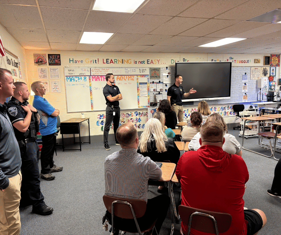 A Milford police officer in uniform speaks with hand gestures to a group of staff members gathered in a hallway or classroom.