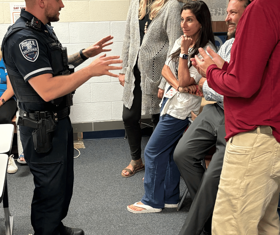A Milford police officer in uniform speaks with hand gestures to a group of staff members gathered in a hallway or classroom.