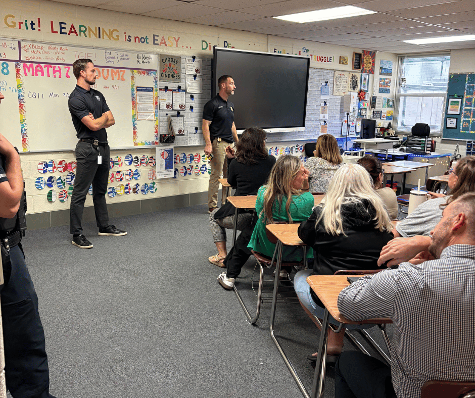 A Milford police officer in uniform speaks with hand gestures to a group of staff members gathered in a hallway or classroom.
