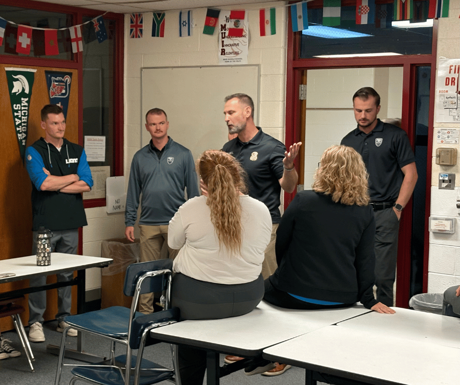 A Milford police officer in uniform speaks with hand gestures to a group of staff members gathered in a hallway or classroom.