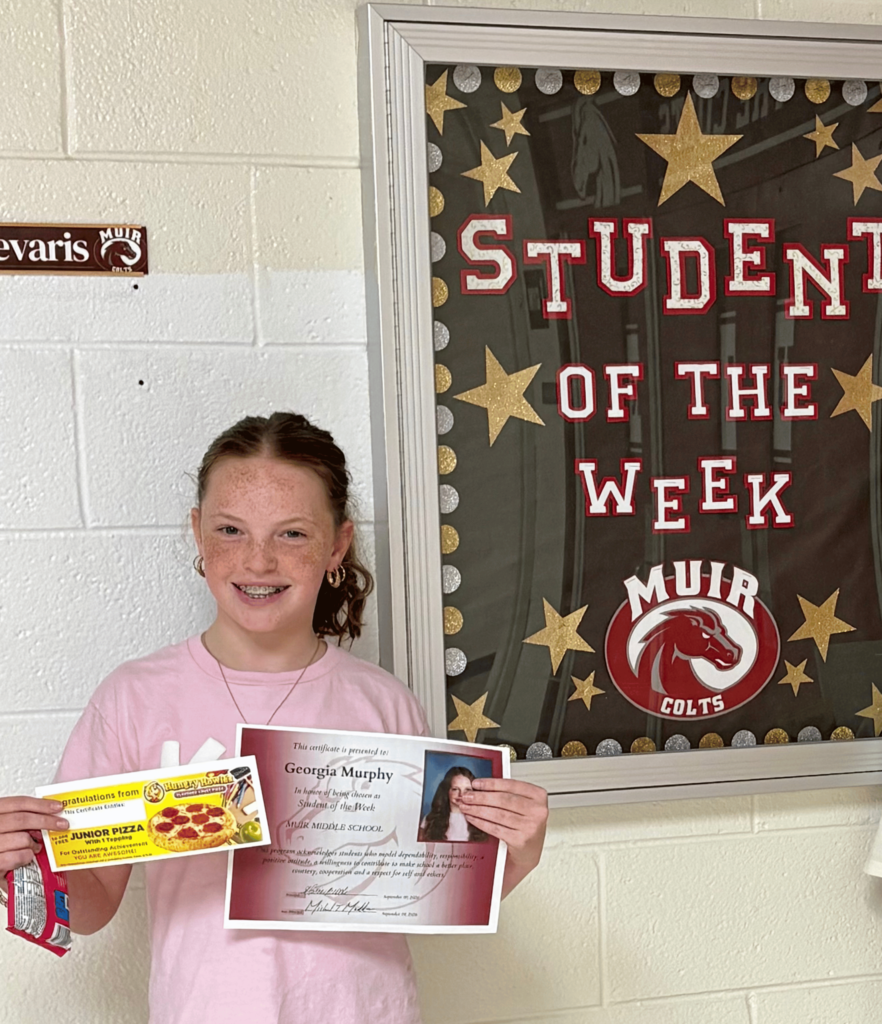Student holding a certificate and candy next to a Student of the Week sign.