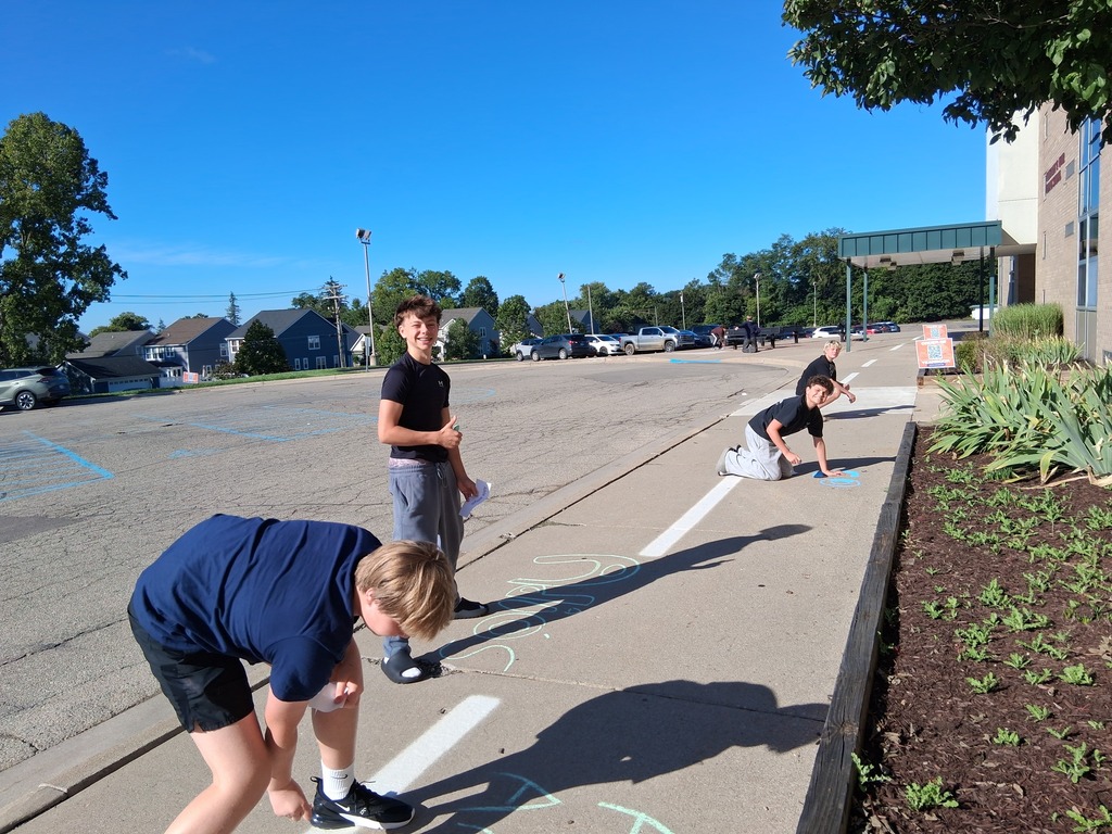 Group of students writing Spanish greetings with sidewalk chalk outside school doors.