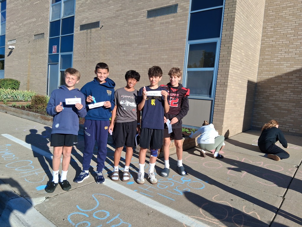 Group of students standing in front of Spanish greetings made  with sidewalk chalk outside school doors.
