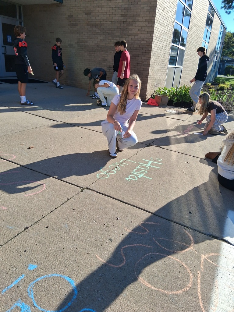 A student kneeling by her Spanish greeting made with sidewalk chalk outside school doors.