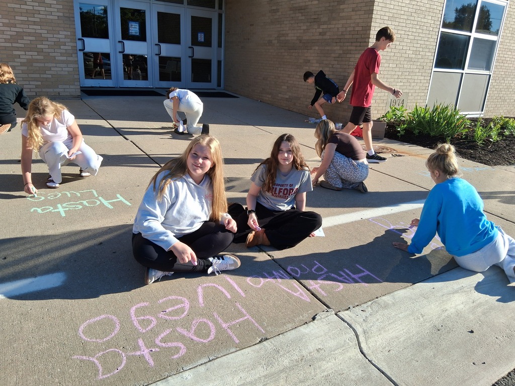 Group of students writing Spanish greetings with sidewalk chalk outside school doors.