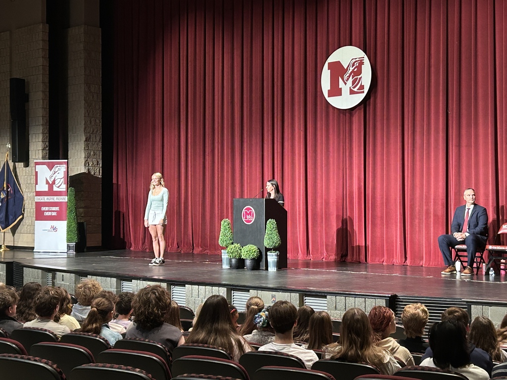 Teen standing on a stage looking at a woman at a podium with a crowd in front of them seated in a theatre