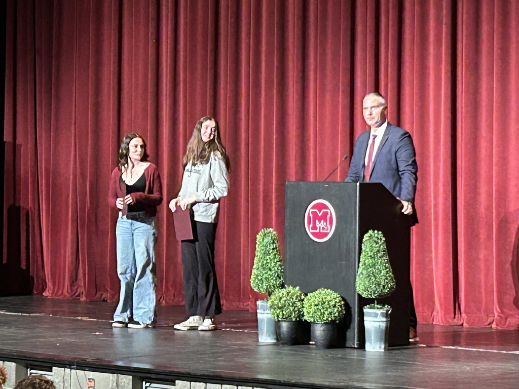 Two teens standing on a stage holding certificates looking at a man at a podium