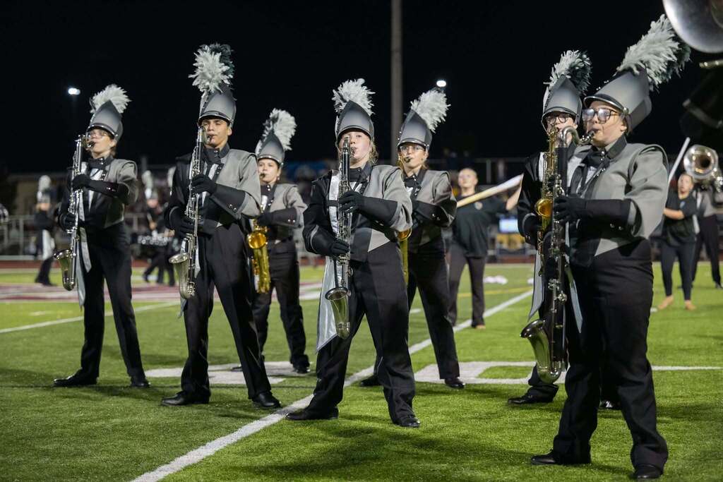 Several band members marching on a football field all wearing black and grey uniformsor practice.