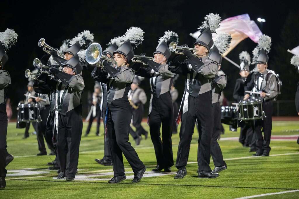 Several band members marching on a football field all wearing black and grey uniformsor practice.