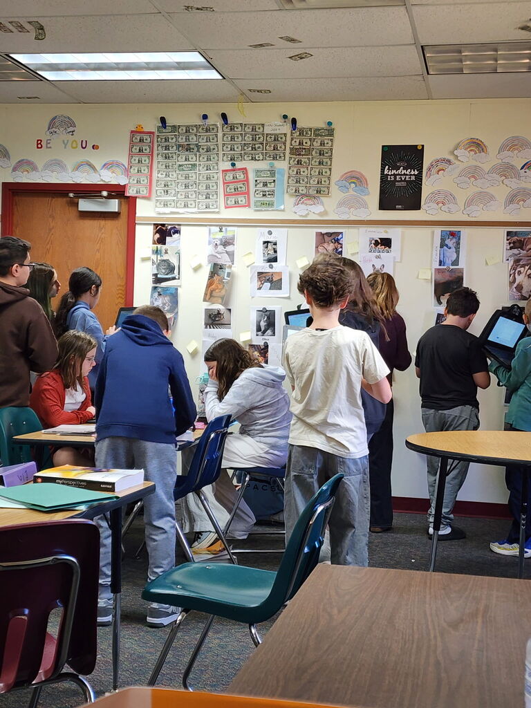 Students gathered in a classroom looking at pictures and posters displayed along a wall board.