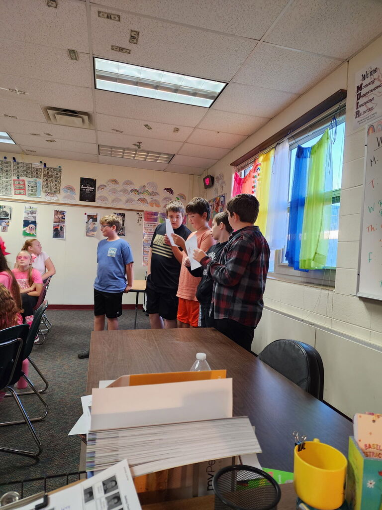 Four students standing at the front of a classroom presenting papers while classmates watch from desks.