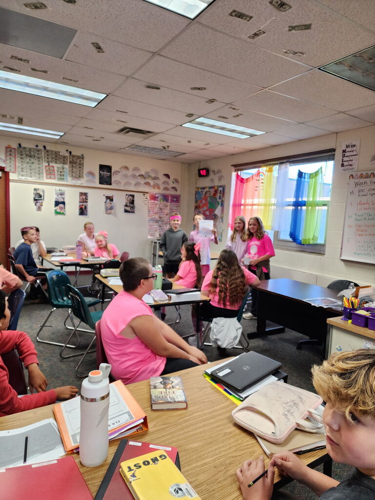 Students wearing pink shirts sit at classroom desks facing a group presenting at the front of the room during a school lesson.