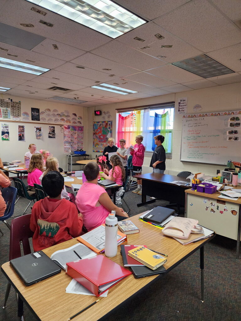 A classroom view from behind student desks as a group of four students presents a project at the front of the room.
