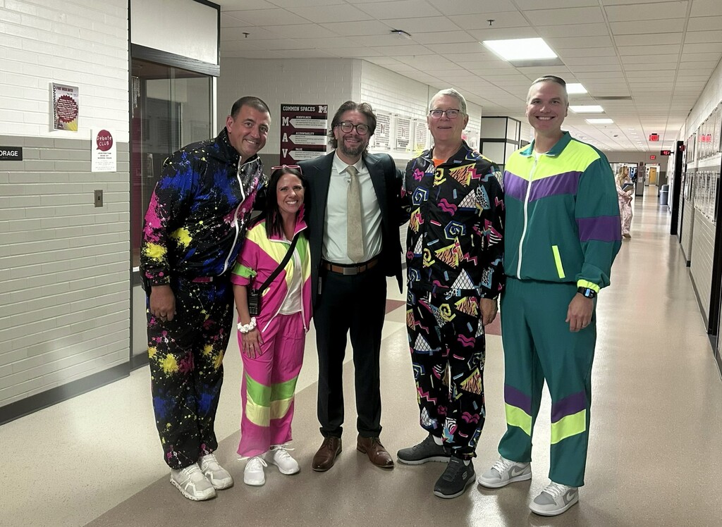 4 men and one woman smiling and posing for a photo in a school hallway. 4 of them are in bright colored track suits. One of them is in a suit.