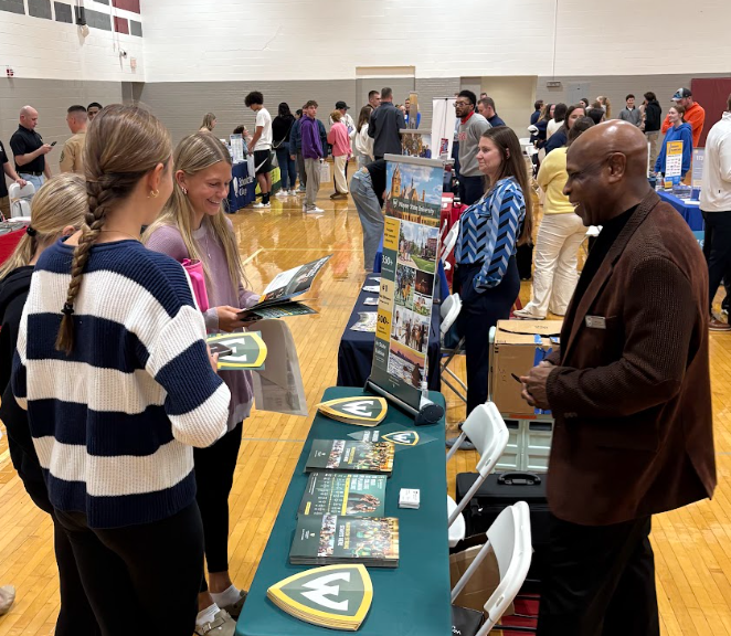 Man talking to two girls at a table.. with dozens of other people wandering around a gymnasium