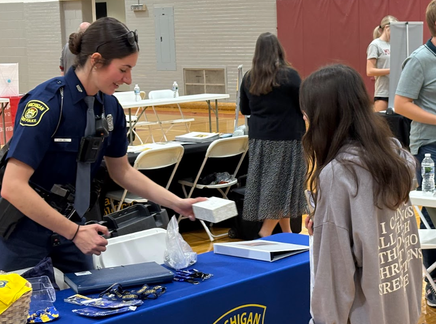 Female police officer talking to a girl at a table.. with other people wandering around a gymnasium behind them