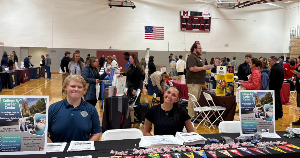 Boy and girl sitting at a table smiling & posing... with dozens of other people wandering around a gymnasium