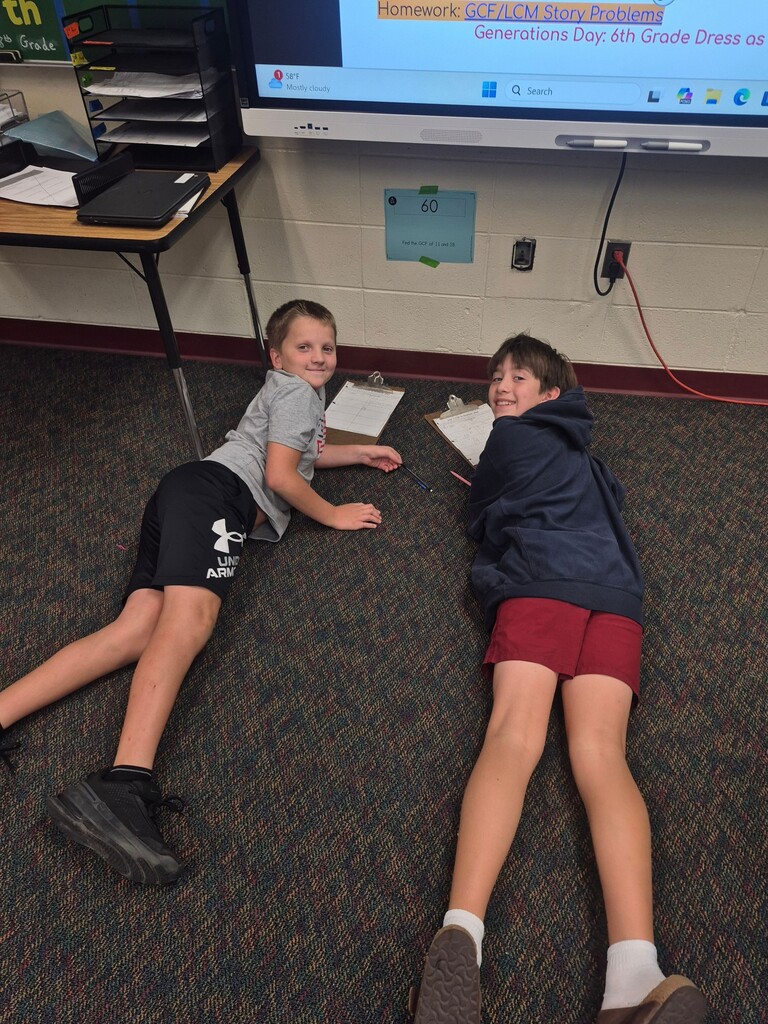 Two students lie on the classroom carpet with clipboards, smiling toward the camera below a digital presentation board.