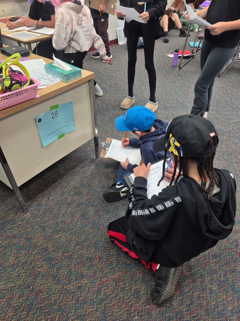 Two students sit on the classroom floor writing on clipboards while classmates stand nearby during a lesson.