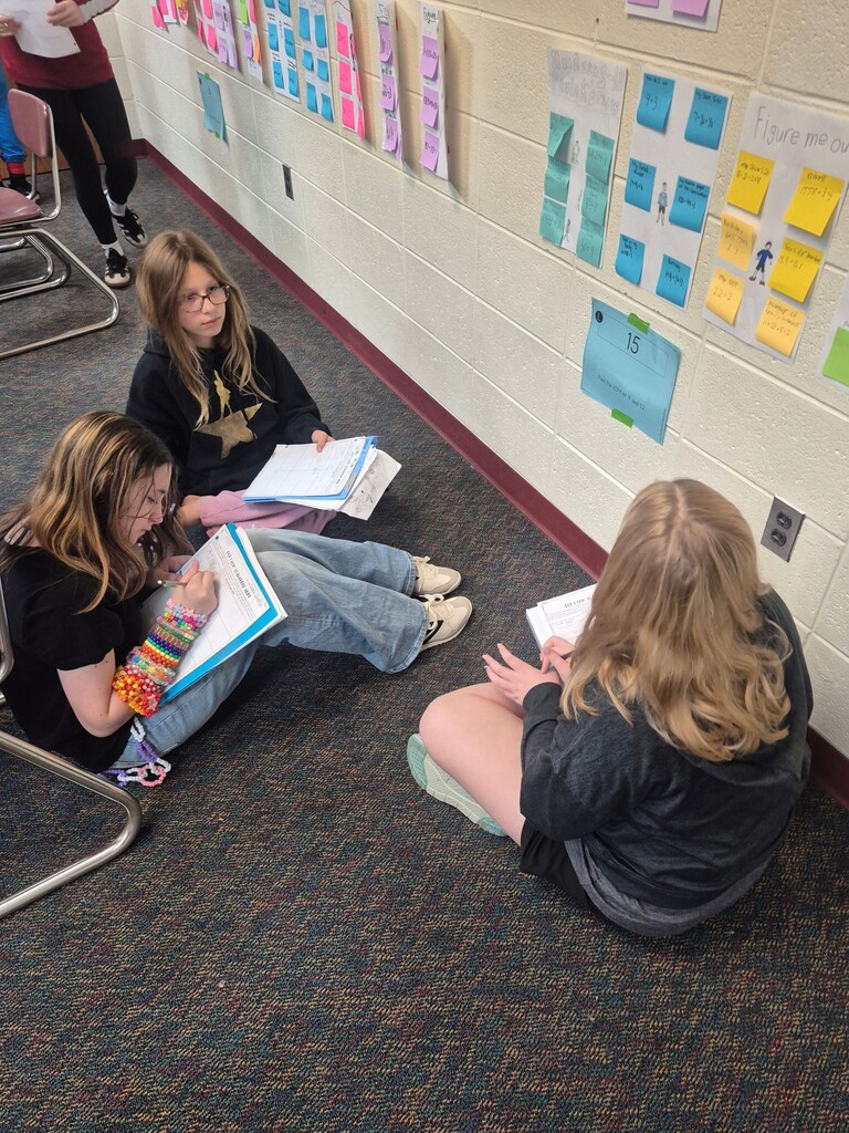 Three students sit in a circle on the classroom carpet writing on clipboards next to a wall activity board.