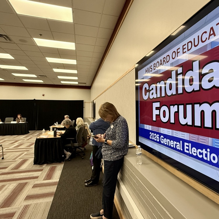 photo of video monitor in foreground that reads “candidate forum” with a conference room full of chairs in the background