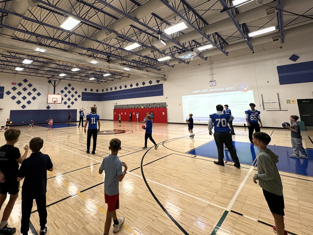 Several Football players in jerseys playing a ball game in a gym with younger students
