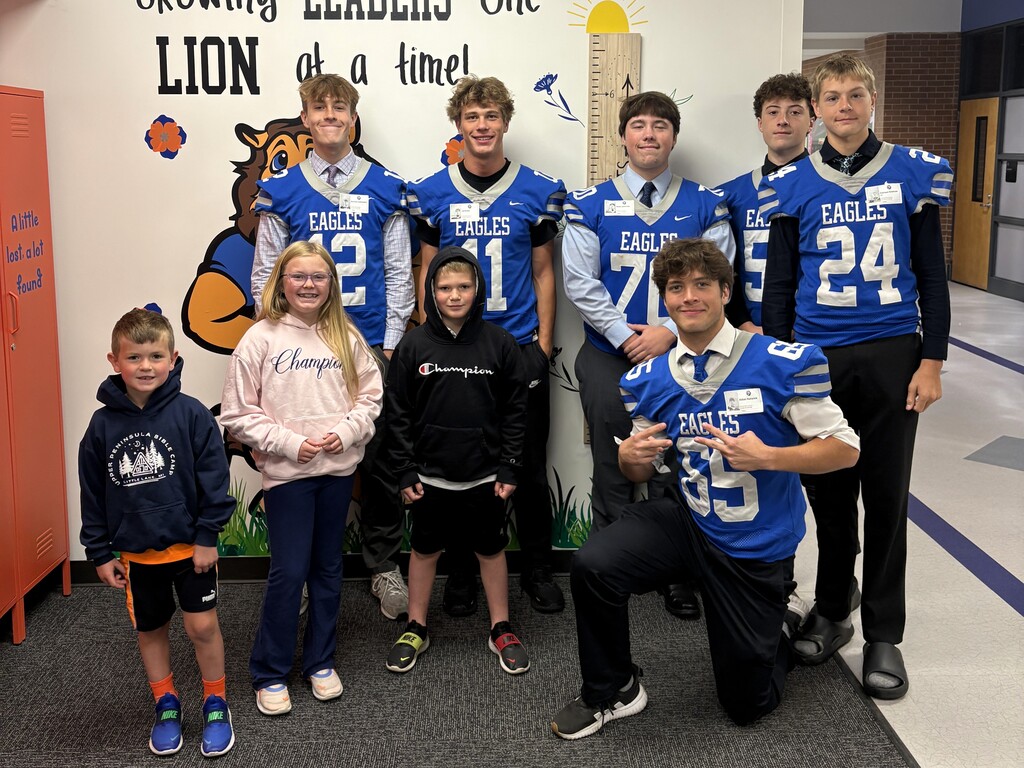 Several Football players in jerseys posing & smiling with younger students for a photo