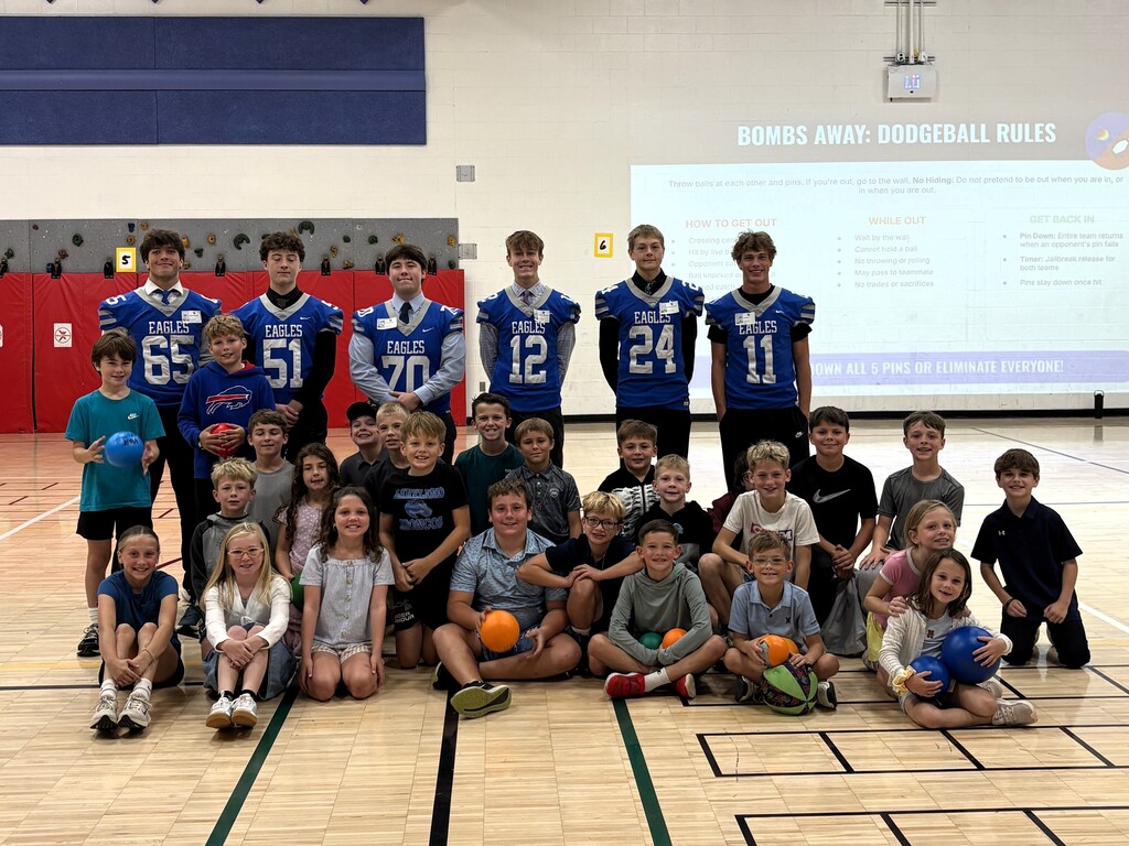 Several Football players in jerseys posing & smiling with younger students for a photo