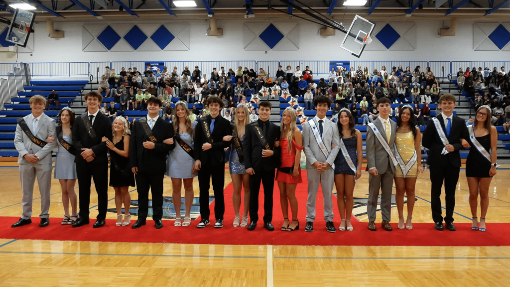 Several male & female students dressed up wearing sashes on the gym floor during an assembly