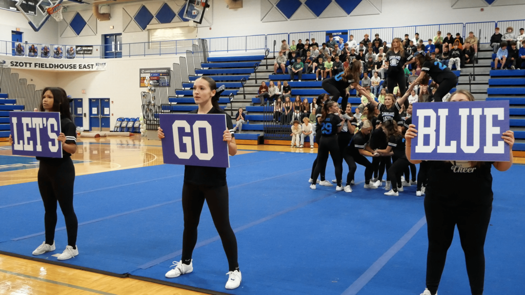 Female students doing cheer routine on gym floor while holding lets go blue signs