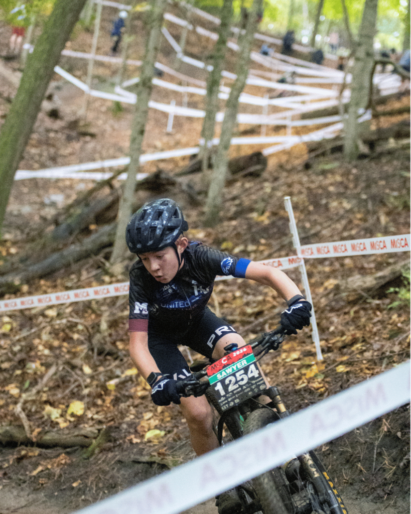 Close-up action shot of a youth mountain biker wearing helmet and gloves focused on navigating a curve on a wooded trail during a race.   