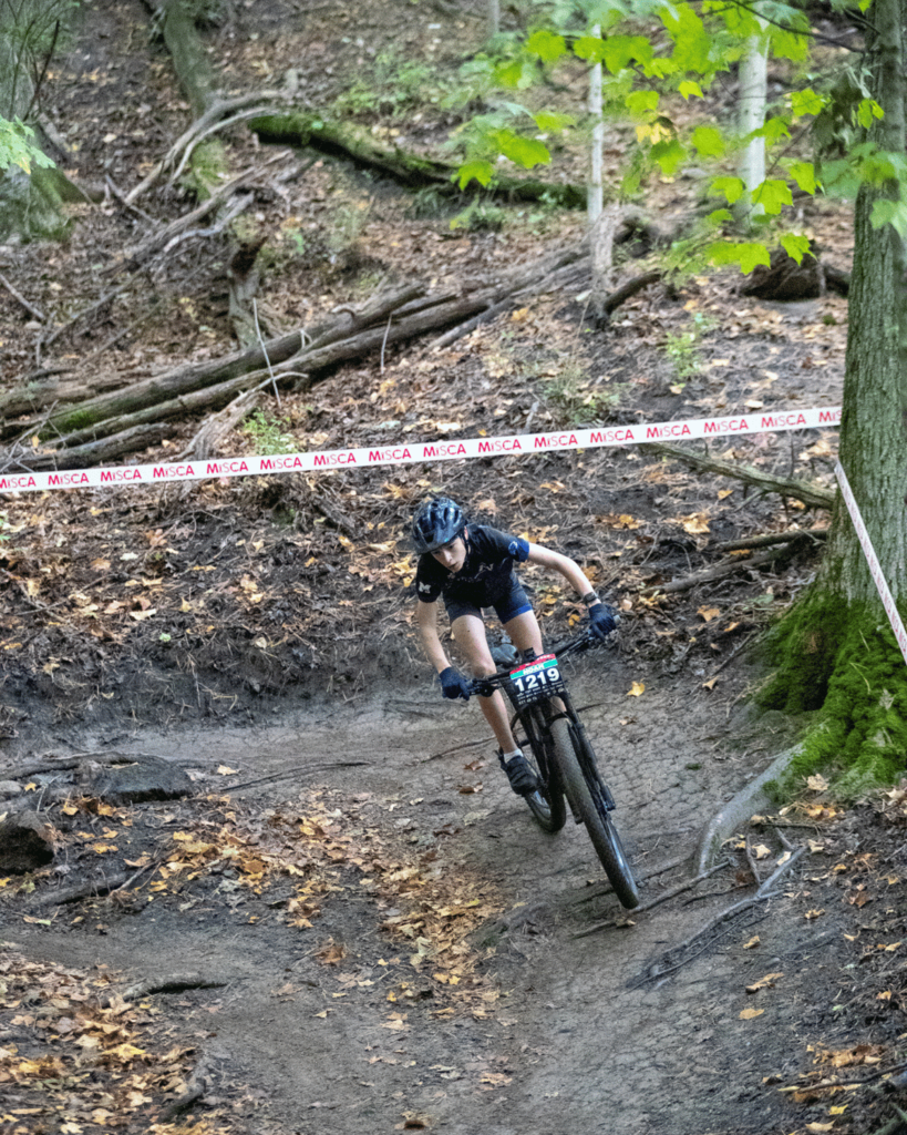 A youth mountain biker wearing a helmet and riding jersey leans into a sharp turn while descending a leaf-covered dirt trail lined with spectator tape.   