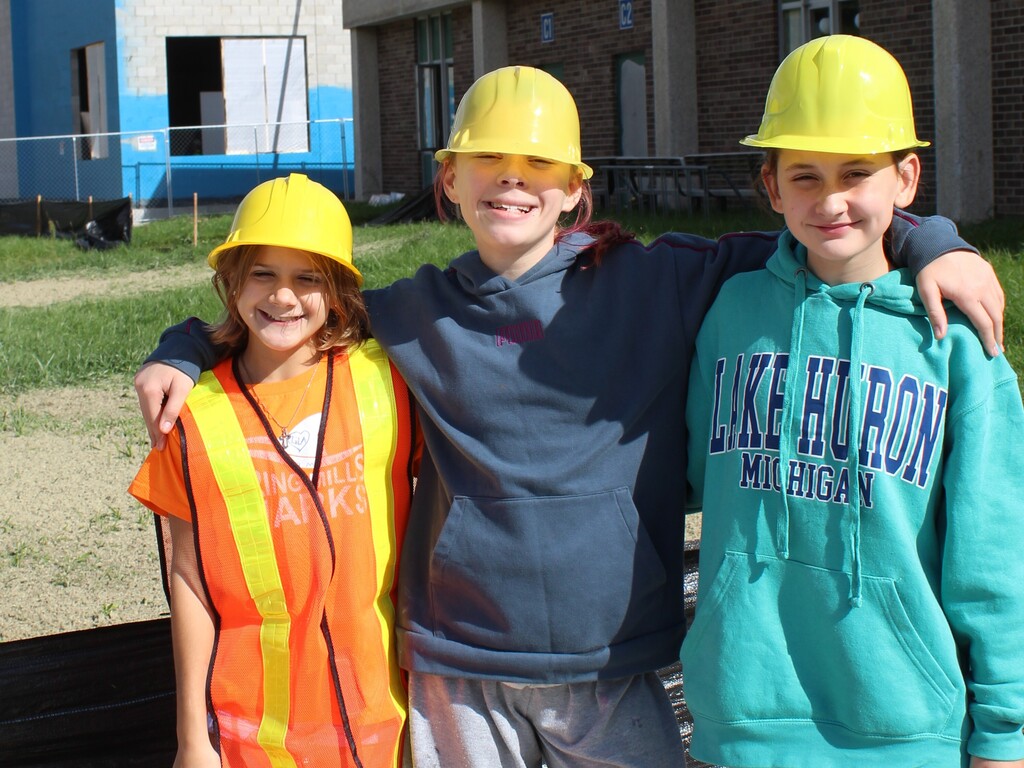 3 children smiling and posing for a photo outside a school wearing construction gear.