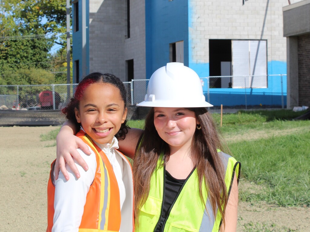 a pair of children smiling and posing for a photo outside a school wearing construction gear.