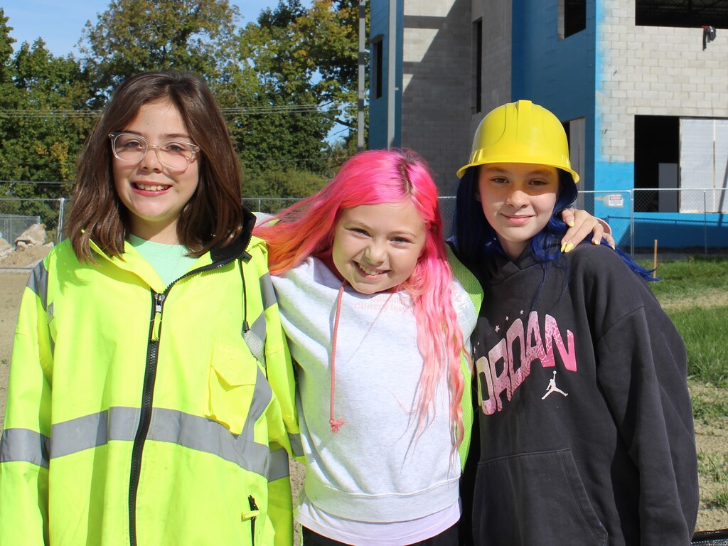 3 children smiling and posing for a photo outside a school wearing construction gear.