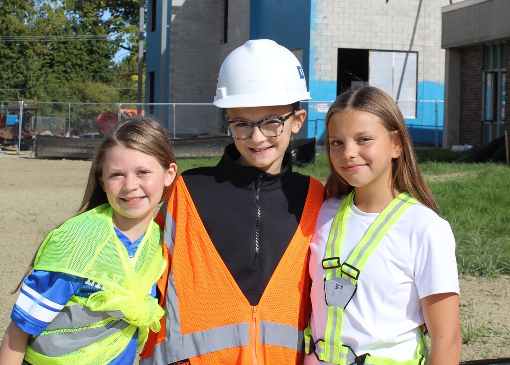 3 children smiling and posing for a photo outside a school wearing construction gear.