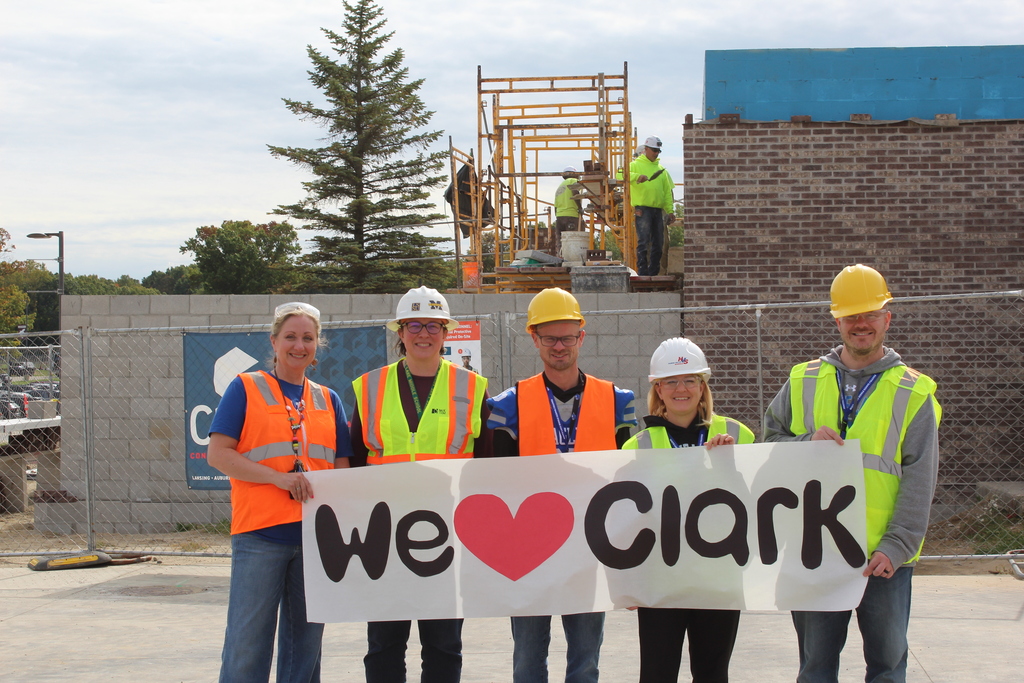 5 adults smiling and posing for a photo outside a school wearing construction gear and holding a sign that reads " We love clark"