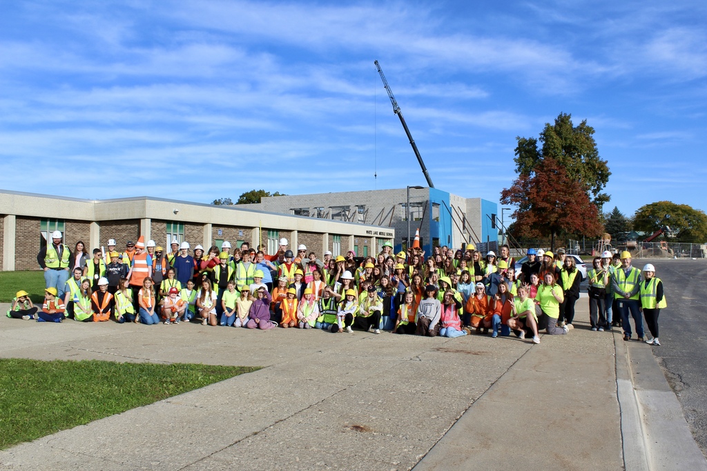 Dozens of students and teachers pose for a group photo in front of a school while construction crews work in the background