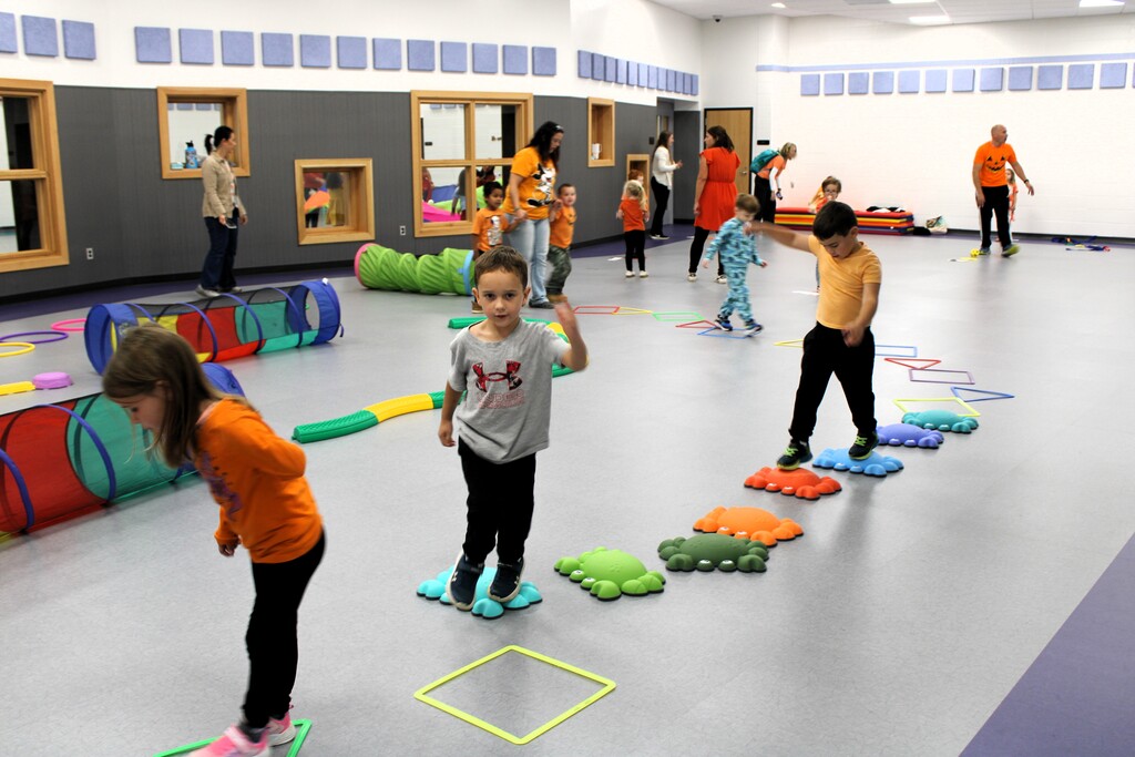 Several kids stepping on cushioned toys in a playroom while other kids & adults do the same int he background
