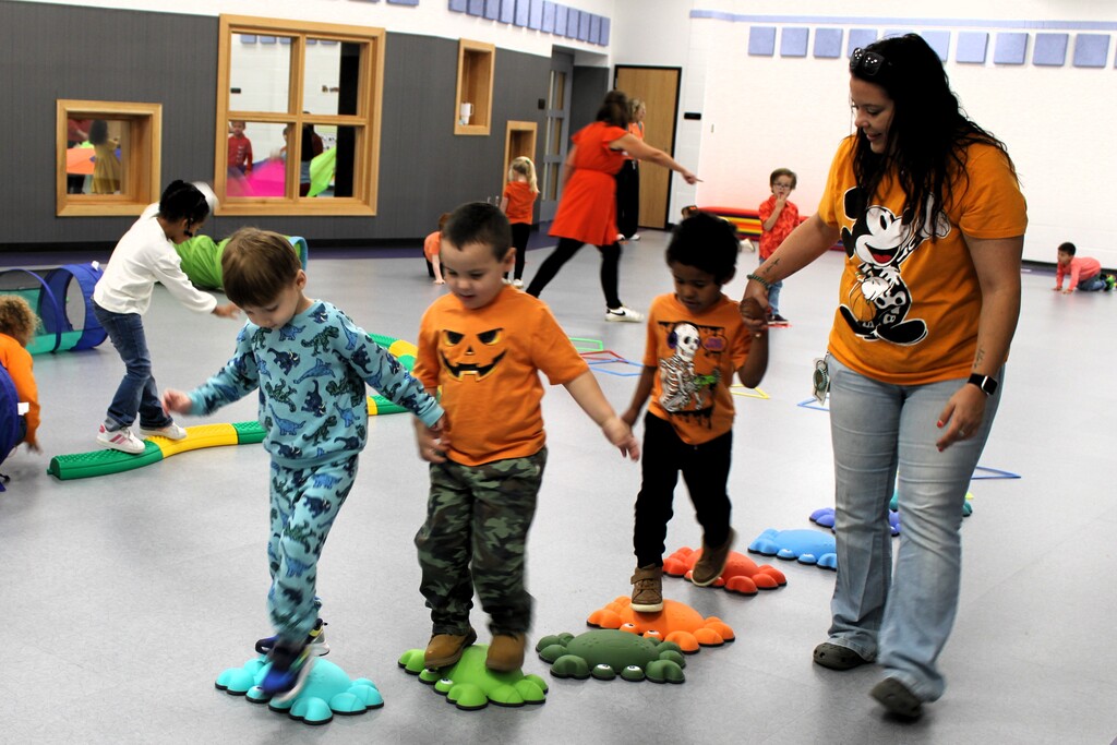 Three kids walking on cushy items on the floor in room while an adult holds one of their hands to assist