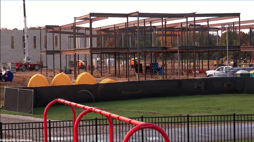 Photo of construction site of a large building with girder framing and a brick building in the background