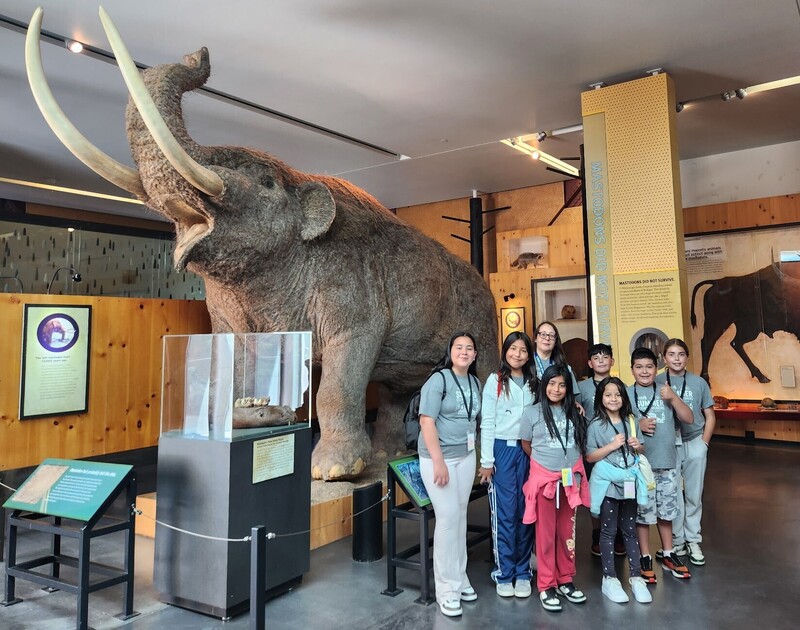 Photo of several students people posing for a photo  standing in front of a mastadon