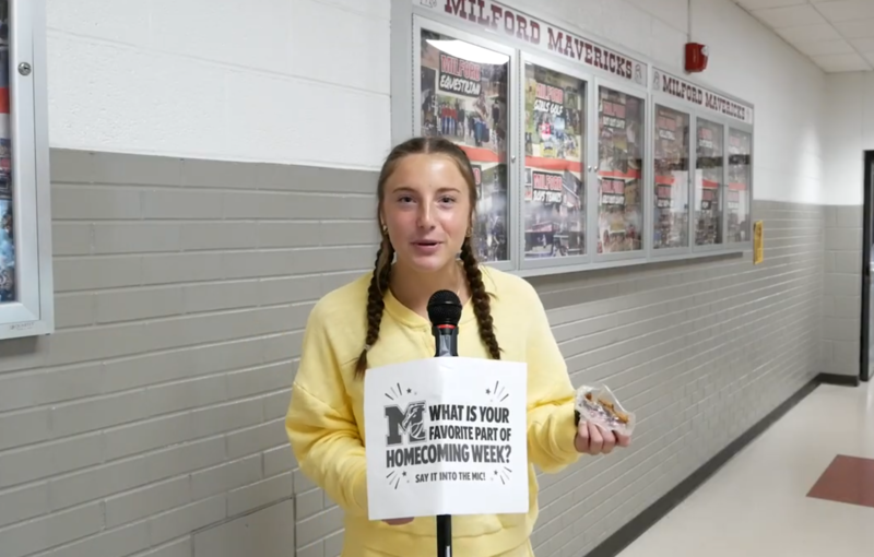 A girl standing in front of a microphone that reads "what's your favorite part of homecoming week?" written on the mic stand