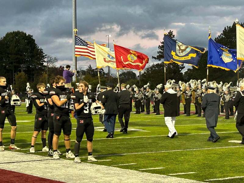 Football players lined up on the sideline of a football field with hands over hearts with color guard walking in the background on football field