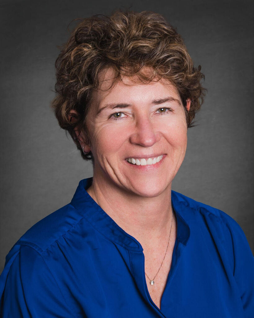 A woman with short, curly brown hair, wearing a blue top and a necklace. She smiles, and her teeth are visible.